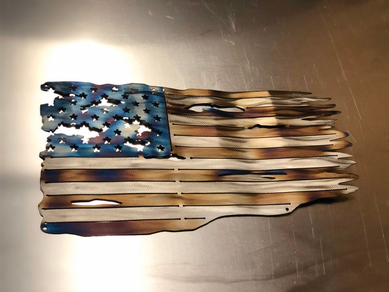 May include: A distressed American flag made of metal with a blue field of stars and red and white stripes. The flag has a weathered, burnt look, giving it a rustic appearance.