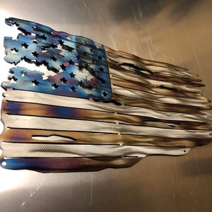 May include: A distressed American flag made of metal with a blue, white, and brown color scheme. The flag is slightly wavy and has a distressed look, with some of the stars and stripes missing.