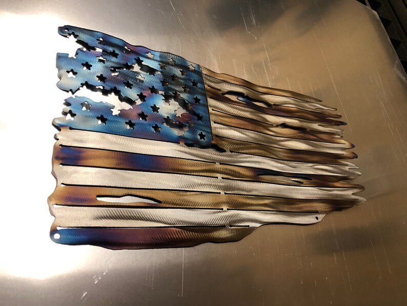 May include: A distressed American flag made of metal with a blue, white, and brown color scheme. The flag is slightly wavy and has a distressed look, with some of the stars and stripes missing.
