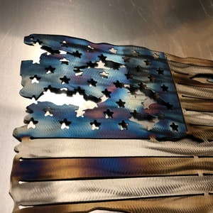 May include: A distressed American flag made of metal with a blue field of stars and red and white stripes. The flag is weathered and has a rustic look.