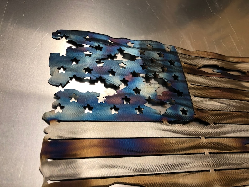 May include: A distressed American flag made of metal with a blue field of stars and red and white stripes. The flag is weathered and has a rustic look.