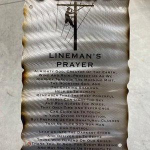 May include: A metal sign with a rustic, weathered look featuring a silhouette of a lineman working on a power line. The sign includes a prayer for linemen, asking for protection and strength. The text reads: "Lineman's Prayer Almighty God, Creator of the Earth, Wind and Rain, Protect us as we journey through the morning mist, the noontime sun, and the evening shadows. Help us to always remember that the most powerful energy can light the sky and run across the wires, that only time and experience can guide us to touch in your divine intervention, but prepare us for unnatural clashes that neither God nor man can control. Lead us into the darkest storm and deliver us to serve those who depend on our skills. Thank you, O' God, for every blessing and may you give us strength to reach for the stars."