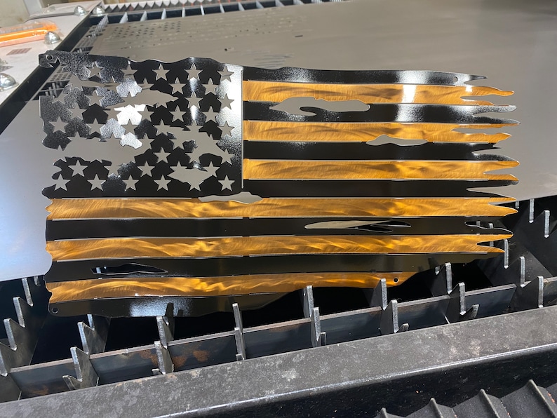 May include: Metal American flag with a distressed design. The flag features cut-out stars and stripes in black and gold. The flag is made of metal and has a weathered appearance.