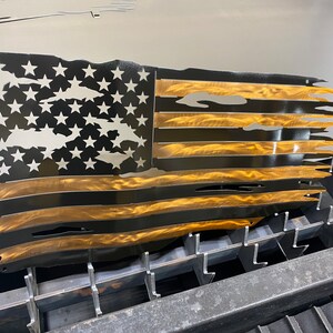 May include: A metal wall art piece depicting a distressed American flag. The flag features black stripes and a gold-colored section, with white stars on a black background. The edges are torn, giving it a weathered appearance.