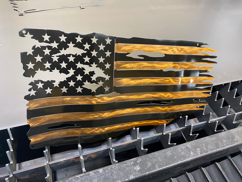 May include: A metal wall art piece depicting a distressed American flag. The flag features black stripes and a gold-colored section, with white stars on a black background. The edges are torn, giving it a weathered appearance.