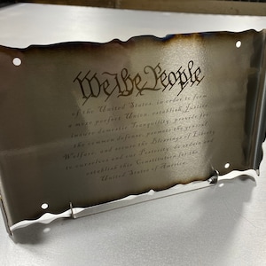 May include: A metal sign with a distressed, aged look, featuring the text "We the People" and the first paragraph of the US Constitution. The sign has a decorative, scroll-like border and is mounted on a stand.