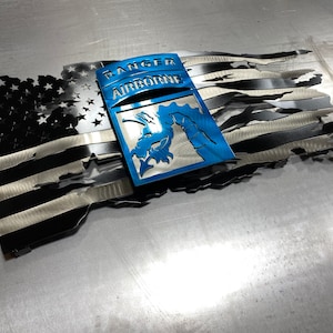 May include: A distressed metal American flag with a blue metal patch that reads "Ranger Airborne" and features a stylized image of a paratrooper.