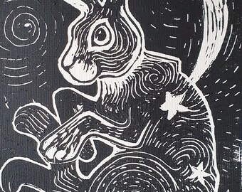 May include: Black and white linocut print of a rabbit with a crescent moon and stars. The rabbit is seated, with detailed linework creating a textured effect. The background is black with swirling patterns.