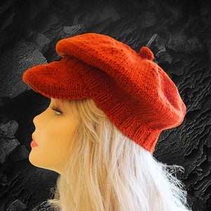 May include: A red knitted newsboy cap with a slightly rounded crown and a brim that curves down on the sides. The cap is made of a soft, textured yarn.