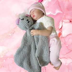 May include: A sleeping baby doll wearing a pink hat with a heart-shaped detail, a white and green shirt, and pink pants. The doll is holding a gray knitted security blanket with a pink and gray bear face.