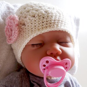 May include: A sleeping baby doll wearing a white crocheted hat with a pink flower accent and a pink pacifier.