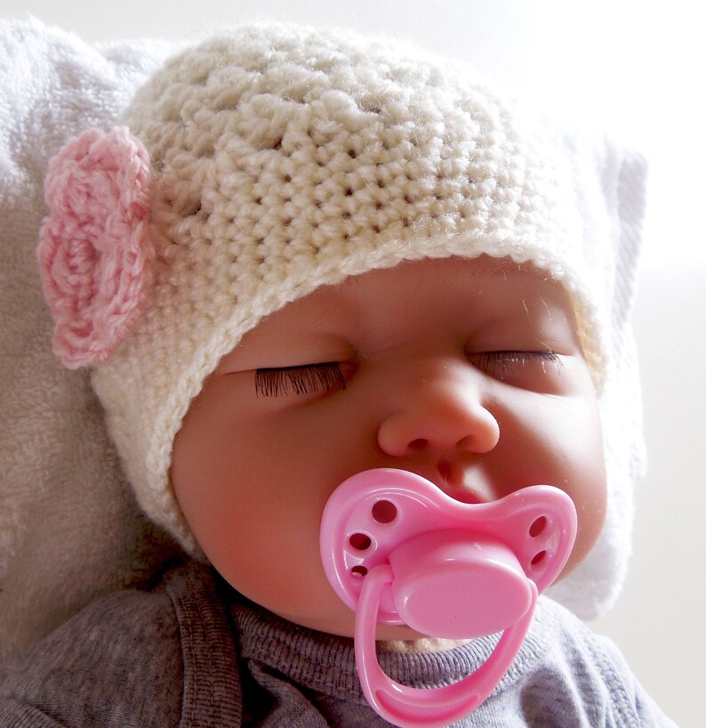 May include: A sleeping baby doll wearing a white crocheted hat with a pink flower accent and a pink pacifier.