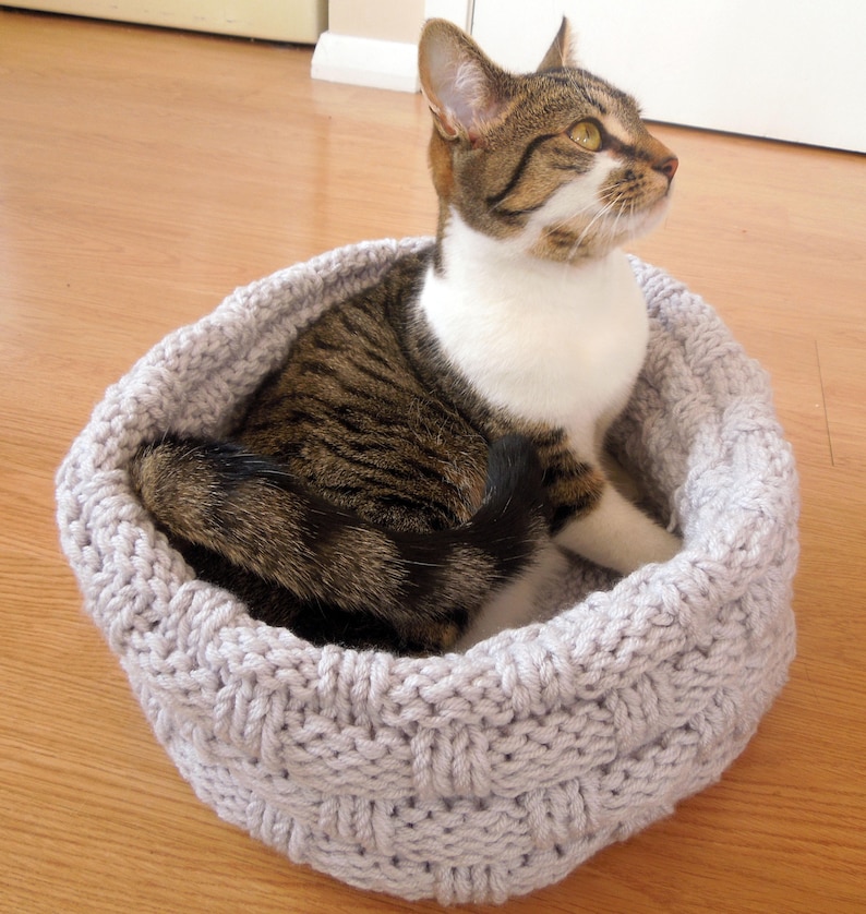May include: A gray knitted cat bed with a tabby cat resting inside. The cat bed is made of a thick, textured yarn and has a round shape.