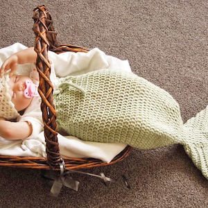 May include: A baby wearing a crocheted mermaid tail and a pink flower hat is lying in a wicker basket. The baby is wearing a pacifier.