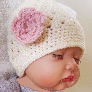 May include: A white crocheted baby hat with a pink crocheted flower on top. The hat is worn by a baby with their eyes closed.