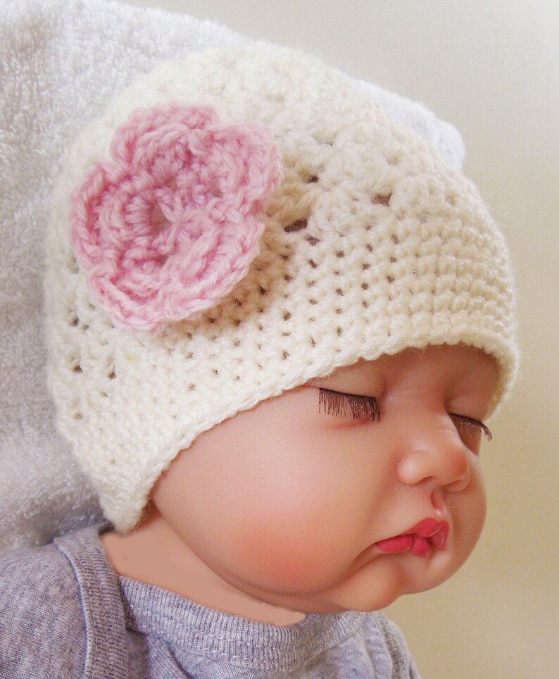 May include: A white crocheted baby hat with a pink crocheted flower on top. The hat is worn by a baby with their eyes closed.