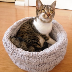 May include: A tabby and white cat resting in a gray knitted cat bed. The cat is looking up and has green eyes.