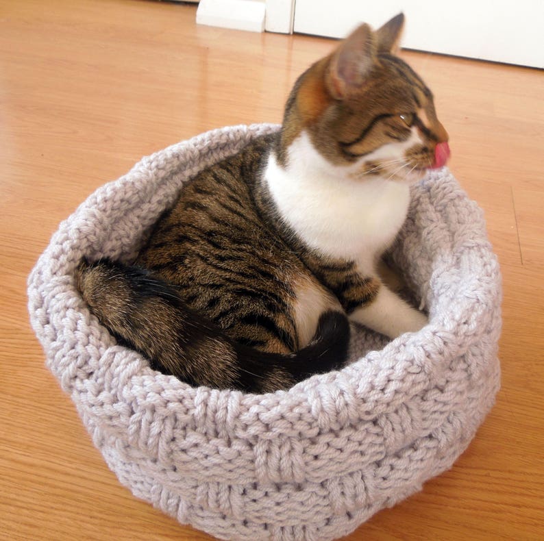 May include: A gray knitted cat bed with a tabby cat resting inside. The cat bed is made with a woven pattern and has a round shape.