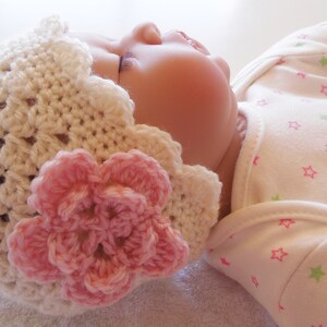 May include: A white crocheted baby hat with a pink flower on top. The hat is made with a delicate stitch pattern and has a scalloped edge.