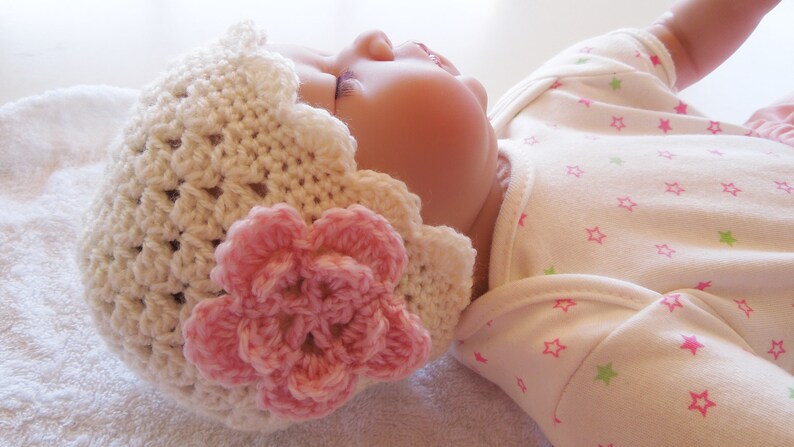 May include: A white crocheted baby hat with a pink flower on top. The hat is made with a delicate stitch pattern and has a scalloped edge.