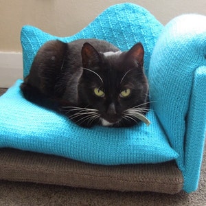 May include: A black cat with white markings is lying on a blue knitted cat bed. The bed has a brown base and a rolled armrest.