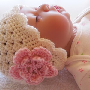 May include: A white crocheted baby hat with a pink flower. The hat has a scalloped edge and is made with a delicate stitch.