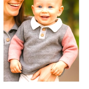May include: A gray and pink sweater with a white collar and wooden buttons. The sweater is worn by a baby with blue eyes. The baby is being held by a person wearing a matching gray sweater.