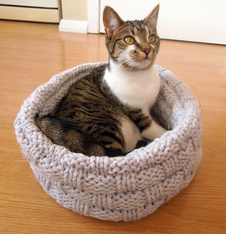 May include: A gray knitted cat bed with a tabby cat sitting inside. The cat bed is made of a thick, woven yarn and has a round shape.