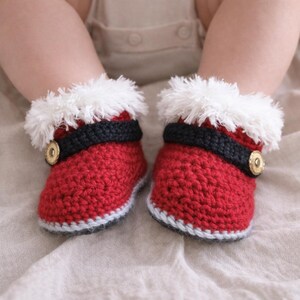 May include: A pair of red crocheted booties featuring a white faux fur trim at the top, a black horizontal band across the middle, and a single gold-toned circular button on the side of each bootie. The base of each bootie has a light gray trim.