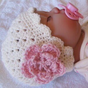 May include: A white crocheted baby hat with a pink flower embellishment. The hat has a scalloped edge and is made with a delicate stitch pattern.