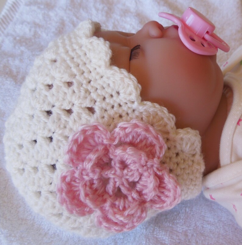May include: A white crocheted baby hat with a pink flower embellishment. The hat has a scalloped edge and is made with a delicate stitch pattern.