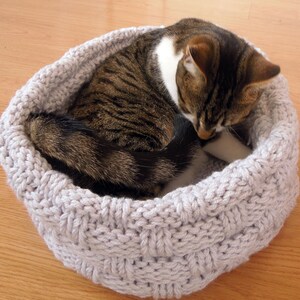 May include: A gray knitted cat bed with a tabby cat curled up inside. The cat bed is made of a soft, cozy material and is perfect for a cat to relax and sleep in.