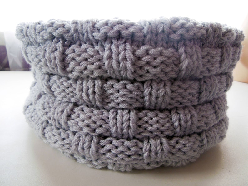 May include: A gray knitted basket with a woven pattern. The basket is made of thick yarn and has a round shape.