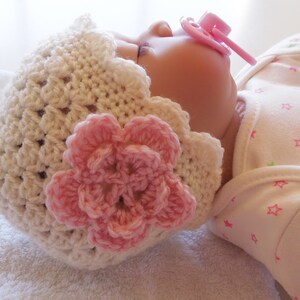 May include: A white crocheted baby hat with a pink flower embellishment. The hat is worn by a baby lying on a white towel.