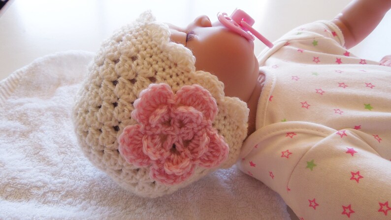 May include: A white crocheted baby hat with a pink flower embellishment. The hat is worn by a baby lying on a white towel.