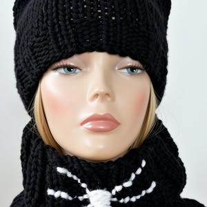 May include: A black chunky knit beanie with two pointed cat ears on top, paired with a matching black chunky knit cowl scarf featuring a white embroidered spider web design on the front. The set is displayed on a mannequin head.