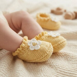 May include: A pair of soft, pale yellow knitted baby booties featuring a ruffled edge and a small, white crocheted flower appliqué with a yellow center on the top of each shoe. The booties are crafted with a textured knit pattern.