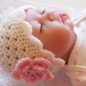 May include: A white crocheted baby hat with a pink flower embellishment. The hat is worn by a sleeping baby with a white onesie with pink and green stars.