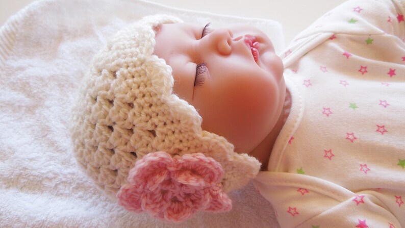 May include: A white crocheted baby hat with a pink flower embellishment. The hat is worn by a sleeping baby with a white onesie with pink and green stars.