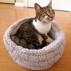 May include: A tabby cat with white markings rests in a gray knitted cat bed. The bed is made of thick, woven yarn and has a round shape.