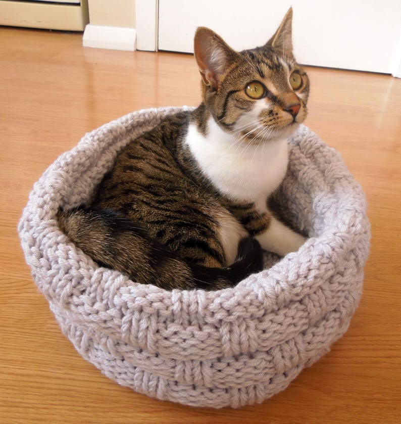 May include: A tabby cat with white markings rests in a gray knitted cat bed. The bed is made of thick, woven yarn and has a round shape.