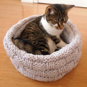 May include: A gray knitted cat bed with a tabby cat resting inside. The cat bed is round and has a woven pattern.