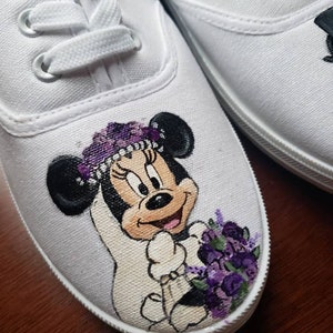 May include: White canvas shoes with a hand-painted design of Minnie Mouse dressed as a bride. She is wearing a white dress, a purple flower crown, and holding a purple bouquet.