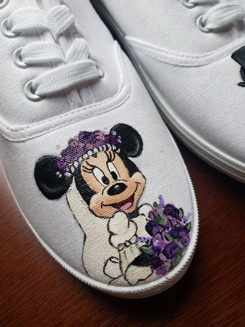 May include: White canvas shoes with a hand-painted design of Minnie Mouse dressed as a bride. She is wearing a white dress, a purple flower crown, and holding a purple bouquet.