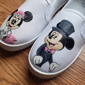May include: A pair of white canvas slip-on shoes with hand-painted images of Mickey Mouse and Minnie Mouse dressed in wedding attire. Mickey Mouse is wearing a tuxedo and a top hat, while Minnie Mouse is wearing a white dress and a veil.