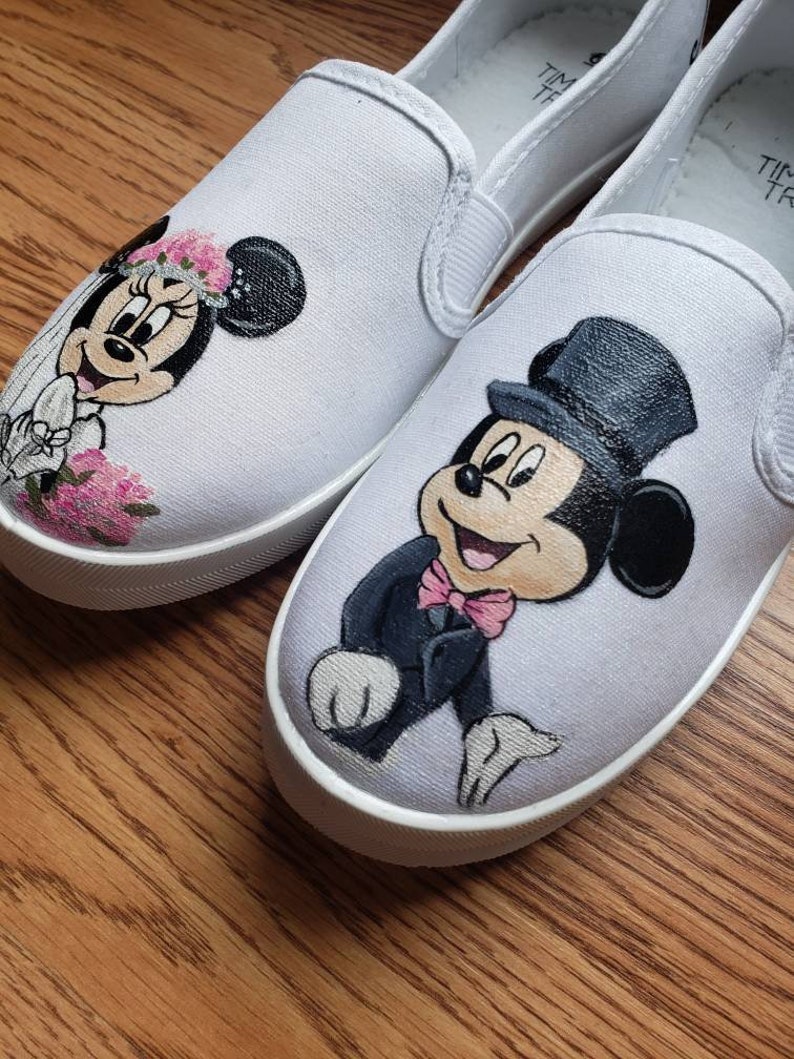 May include: A pair of white canvas slip-on shoes with hand-painted images of Mickey Mouse and Minnie Mouse dressed in wedding attire. Mickey Mouse is wearing a tuxedo and a top hat, while Minnie Mouse is wearing a white dress and a veil.