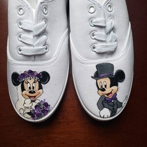 May include: A pair of white canvas shoes with hand-painted images of Mickey and Minnie Mouse dressed in wedding attire. Minnie Mouse is wearing a white dress with a purple flower crown and a bouquet of purple flowers. Mickey Mouse is wearing a black tuxedo with a purple bow tie and a top hat.