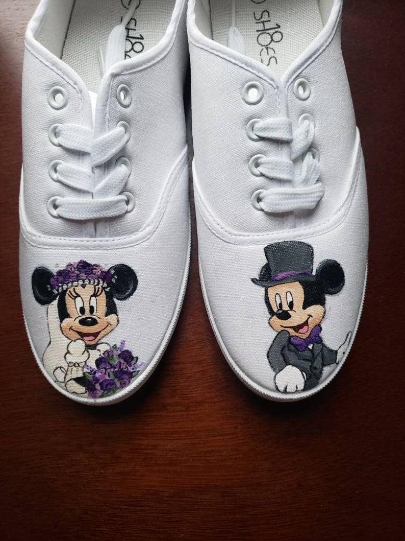 May include: A pair of white canvas shoes with hand-painted images of Mickey and Minnie Mouse dressed in wedding attire. Minnie Mouse is wearing a white dress with a purple flower crown and a bouquet of purple flowers. Mickey Mouse is wearing a black tuxedo with a purple bow tie and a top hat.