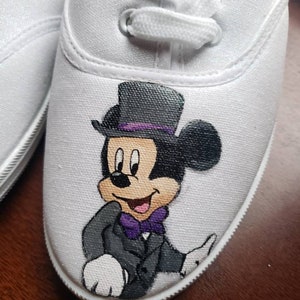 May include: White canvas shoes with a hand-painted design of Mickey Mouse wearing a tuxedo and a top hat. The design is in black, white, purple, and gray.