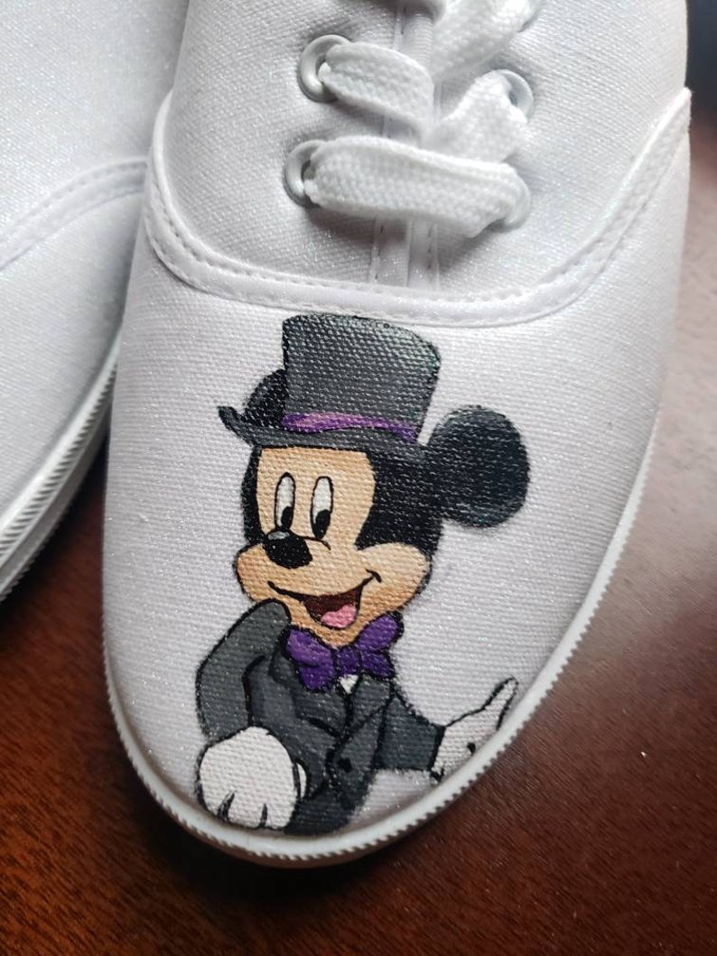 May include: White canvas shoes with a hand-painted design of Mickey Mouse wearing a tuxedo and a top hat. The design is in black, white, purple, and gray.